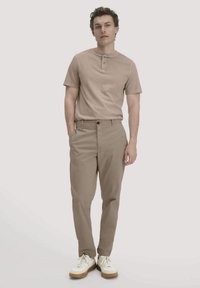 Mann in beigem kurzärmligem Henley-Shirt, passenden beigen Hosen und weißen Sneakers, steht mit einer Hand in der Tasche.