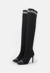 Bottes noires en tricot montant jusqu'à la cuisse avec un motif texturé, pointe noire pointue, talon haut et bande logo blanche contrastante en haut.