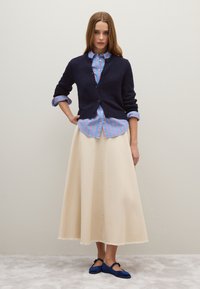 Cardigan lavorato a maglia blu navy con chiusure a bottone sopra una maglietta a righe blu e rosa; gonna maxi color crema con frange; scarpe blu senza lacci.