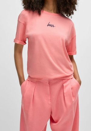 Kobieta w koralowym różowym T-shircie z podwiniętymi rękawami i logo "Boss" oraz dopasowanych plisowanych spodniach, z rękami w kieszeniach, stojąca na jednolitym tle.