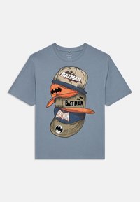 Zila kokvilnas T-krekls ar grafiku, kurā attēloti sakrauti cepures ar tekstu "BATMAN", dažādu stilu un krāsu cepurēm, tostarp oranžiem un bēšiem akcentiem.