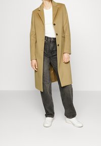 Cappotto in misto lana beige con revers a lancia, chiusura a un bottone, tasche laterali; abbinato a jeans neri a vita alta e sneakers bianche.