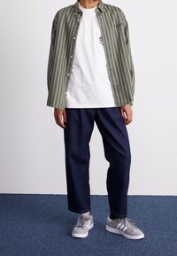 Męski strój składający się z zielonej koszuli w paski na guziki, białej koszulki z kieszenią, ciemnych dżinsów oraz szarych sneakersów z białymi akcentami.