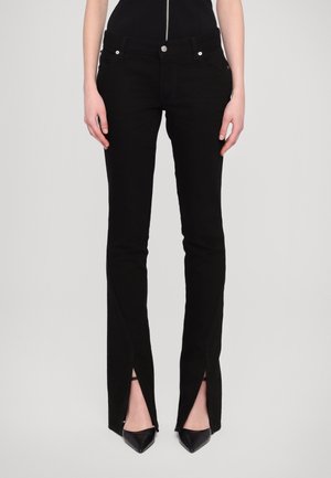 Black denim jeans with a slim fit, featuring front pockets, a zip fly, and side slits at the ankles, paired with pointed black heels.