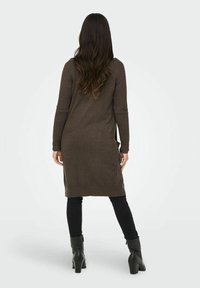 Brązowy, dziergany długi sweter o prostym kroju, z długimi rękawami i rozcięciami po bokach, noszony na czarnych spodniach i czarnych botkach.