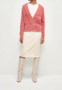 Cardigan en mélange de laine rose avec double boutons dorés et deux poches avant, porté sur une jupe crème et des bottes montantes assorties.