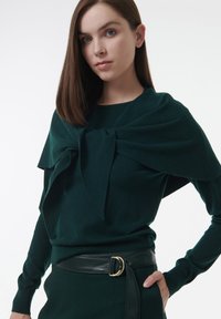 Junge Frau mit glattem braunem Haar, die einen dunkelgrünen Pullover trägt, über den sie ein passendes Tuch über die Schultern gebunden hat, sowie einen schwarzen Gürtel.