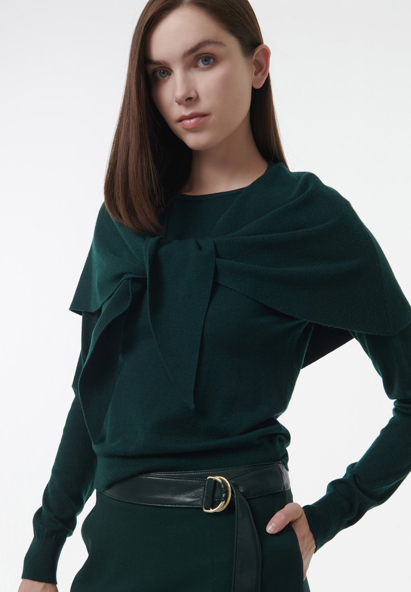 Junge Frau mit glattem braunem Haar, die einen dunkelgrünen Pullover trägt, über den sie ein passendes Tuch über die Schultern gebunden hat, sowie einen schwarzen Gürtel.