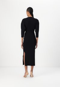 Robe en tricot noir à côtes avec des manches longues et une fente haute. Présente une coupe ajustée, une texture lisse et est associée à des talons nude.