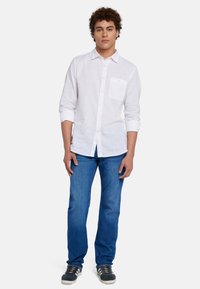 Camicia bianca a maniche lunghe con taschino sul petto, abbinata a jeans blu e sneaker scure. Tessuto semplice, vestibilità rilassata, stile casual.