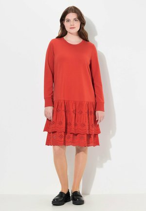 Vestido rojo con mangas largas, parte superior lisa y falda de capas con ojales y patrones florales. Se lleva con zapatos negros, exhibido sobre un fondo blanco.