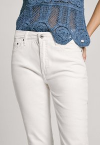 Lys blå hekletrøye med sirkulære detaljer, akkompagnert av hvite denimjeans med glidelås og sidelommer. Jevn tekstur.
