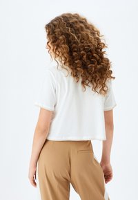Camiseta blanca cortada de manga corta, con un dobladillo texturizado. Combinada con pantalones marrón claro. El fondo es blanco liso.