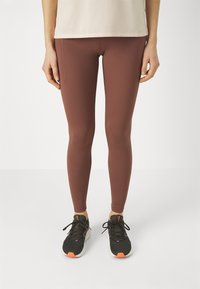 Ben som bär mauve leggings med sidofickor och svarta träningsskor med vita sulor och orangea accenter på en vit bakgrund.