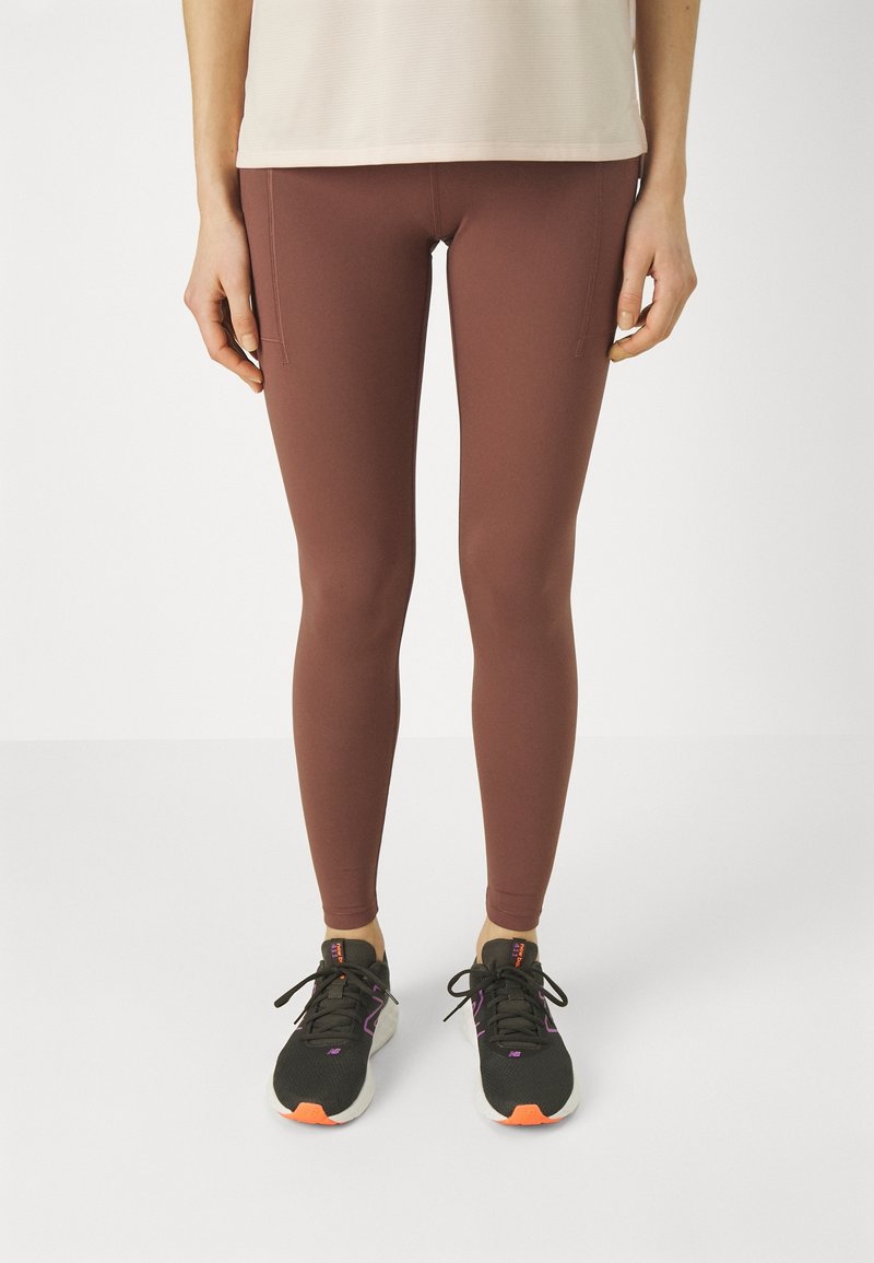 Ben som bär mauve leggings med sidofickor och svarta träningsskor med vita sulor och orangea accenter på en vit bakgrund.