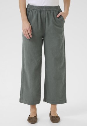 Personne portant un pantalon large gris avec taille élastique et des ballerines marron, une main dans la poche et l'autre reposant sur le côté.