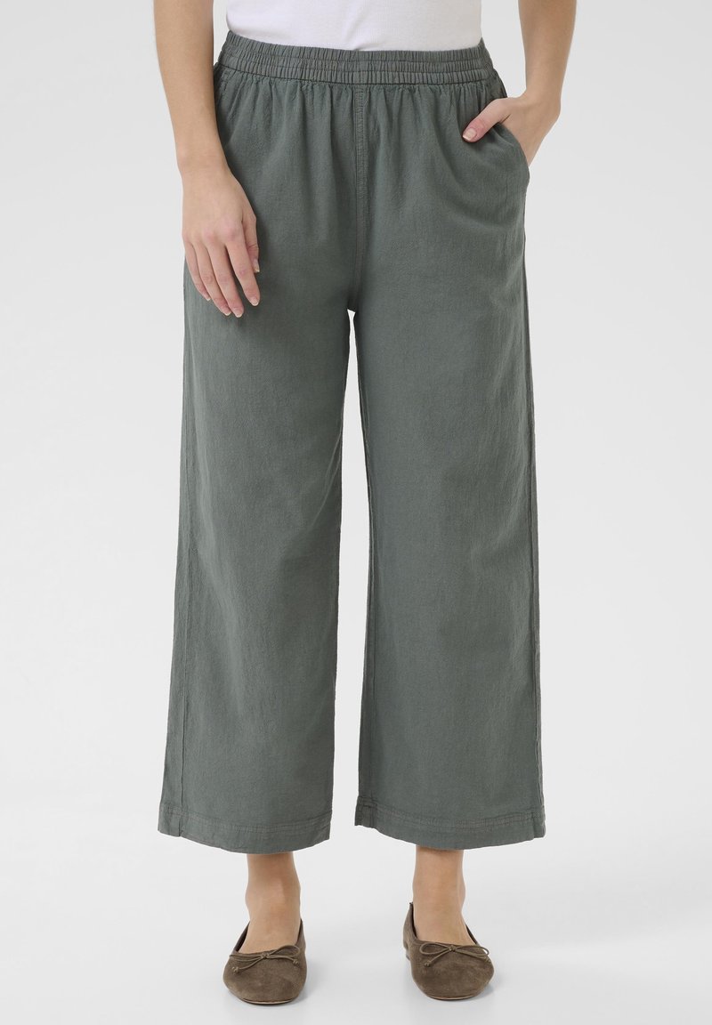 Personne portant un pantalon large gris avec taille élastique et des ballerines marron, une main dans la poche et l'autre reposant sur le côté.