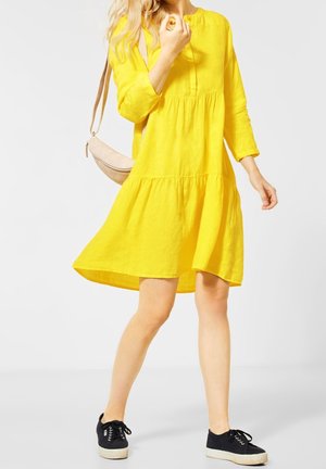 Mujer con un vestido amarillo brillante hasta la rodilla y zapatillas negras, llevando un bolso bandolera beige, caminando sobre un fondo claro.