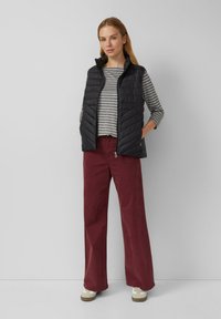 Vrouw staand met handen in de zakken, gekleed in een zwarte gewatteerde vest, gestreepte lange mouw top, brede bordeauxrode broek en witte sneakers.