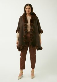 Poncho in lana marrone con collo in pelliccia marrone e dettagli a pom-pom. Indossato sopra un top beige e pantaloni marroni, abbinato a accessori dorati.
