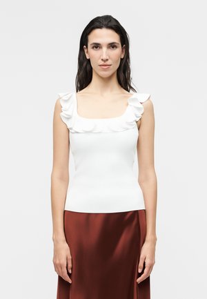 Femme aux longs cheveux foncés portant un haut blanc sans manches avec des épaules à volants et une jupe en satin marron, debout devant un fond uni.