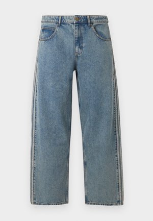 Uomo con jeans azzurri a gamba larga e sneakers beige, in piedi davanti a uno sfondo bianco neutro.
