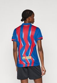 Μπλε και κόκκινη φανέλα με ζιγκ-ζαγκ σχέδια, κοντά μανίκια και ριμπ στο γιακά. Φέρει το λογότυπο του UNHCR σε κίτρινο στο τελείωμα. Μαύρο σορτς.