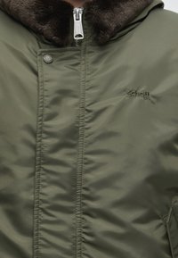 Veste d'hiver verte avec col en fourrure marron, fermeture éclair devant, fermeture à boutons-pression, et petit logo Schott brodé sur la poitrine.