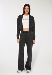 Młoda kobieta w czarnej krótkiej bluzie z kapturem, białej koszulce z grafiką, czarnych spodniach z wysokim stanem i srebrnych trampkach, stoi na białym tle.