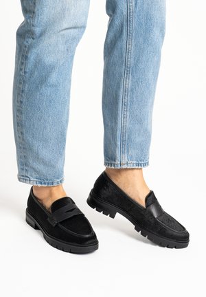 Black leather loafers with chunky soles worn with light blue jeans, shown from mid-calf down against a white background.