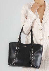 Borsa a tracolla nera in pelle con borchie metalliche lungo i lati, doppie cinghie per le spalle e un accento di logo dorato sul davanti. Texture liscia.