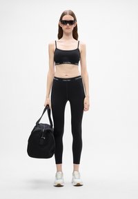 Zwarte sportieve crop top en leggings met Calvin Klein branding, gecombineerd met een elegante zwarte duffel bag en witte sneakers met lichtblauwe accenten.