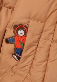 Pufjakas gaiši brūnā krāsā ar flīsa oderi un lāča aplikāciju. Lācis valkā sarkanu kapuci un zilu džinsu, ar zaļo izšuvumu detaļām.