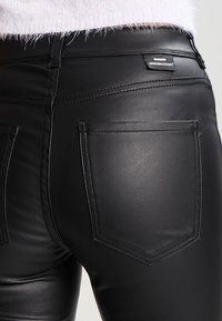 Pantaloni in pelle nera con una texture liscia, due tasche posteriori e cuciture distintive. Presenta un'etichetta del marchio vicino alla vita.