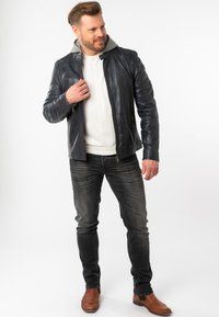 Dunkelblaue Lederjacke mit grauem Kapuzenfutter, taillierter Schnitt, mit Reißverschluss, getragen über einem weißen Hemd und kombiniert mit grauen Jeans und braunen Stiefeln.