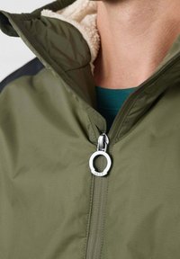Chaqueta verde oliva con cierre delantero con cremallera, que presenta un forro suave de color crema y un tirador circular plateado con la marca "JOTT".