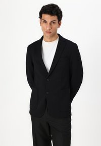 Črn blazer iz gladkega materiala, opremljen z dvema sprednjima žepoma in dvema gumboma, oblečen skupaj z belim majico.