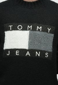 Μαύρο πουλόβερ με ορθογώνιο πλεκτό σχέδιο σε γκρι, ανοιχτό γκρι και λευκά τετράγωνα. Η φράση "TOMMY JEANS" εμφανίζεται έντονα με λευκά γράμματα.