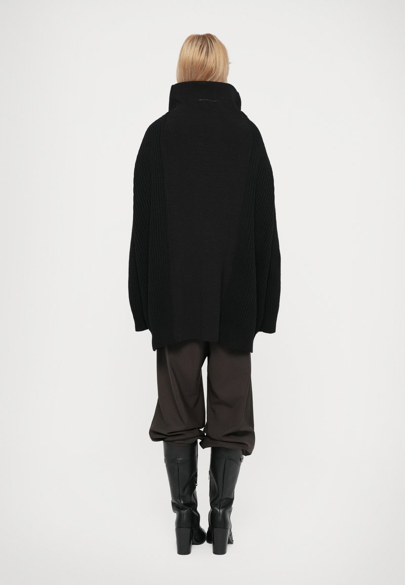 Poncho negro de punto sobredimensionado con cuello alto y textura acanalada, combinado con pantalones negros holgados y botas hasta la rodilla.