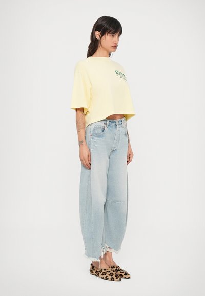 T-shirt jaune court avec texte vert, associée à un jean large déchiré bleu clair et des chaussures plates à motif animal.