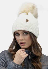 Bonnet tricoté blanc avec un pompon en fausse fourrure sur le dessus, doté d'une petite étiquette en cuir marron. Porté avec une veste zippée grise.