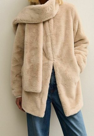 Personne portant un manteau en fausse fourrure beige avec une écharpe assortie, associée à un jean bleu, debout devant un fond uni clair.