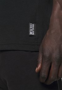 Sort bomulds T-shirt med et synligt logo-tag, der læser "Versace Jeans Couture" på kanten; nærbillede af en hånd mod stoffet.
