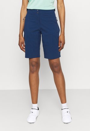 Persoon draagt marineblauwe knielange korte broek, witte sokken en witte sportschoenen, staand met armen ontspannen langs het lichaam.