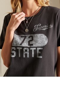 Camiseta negra de manga corta con estampado gráfico gris que incluye "72 STATE" y el logo de "Superdry", cuello redondo y textura suave de algodón.