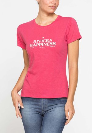 Donna che indossa una t-shirt rosa a maniche corte con il testo "Riviera Happiness" e jeans blu, in piedi con una mano vicino al fianco.