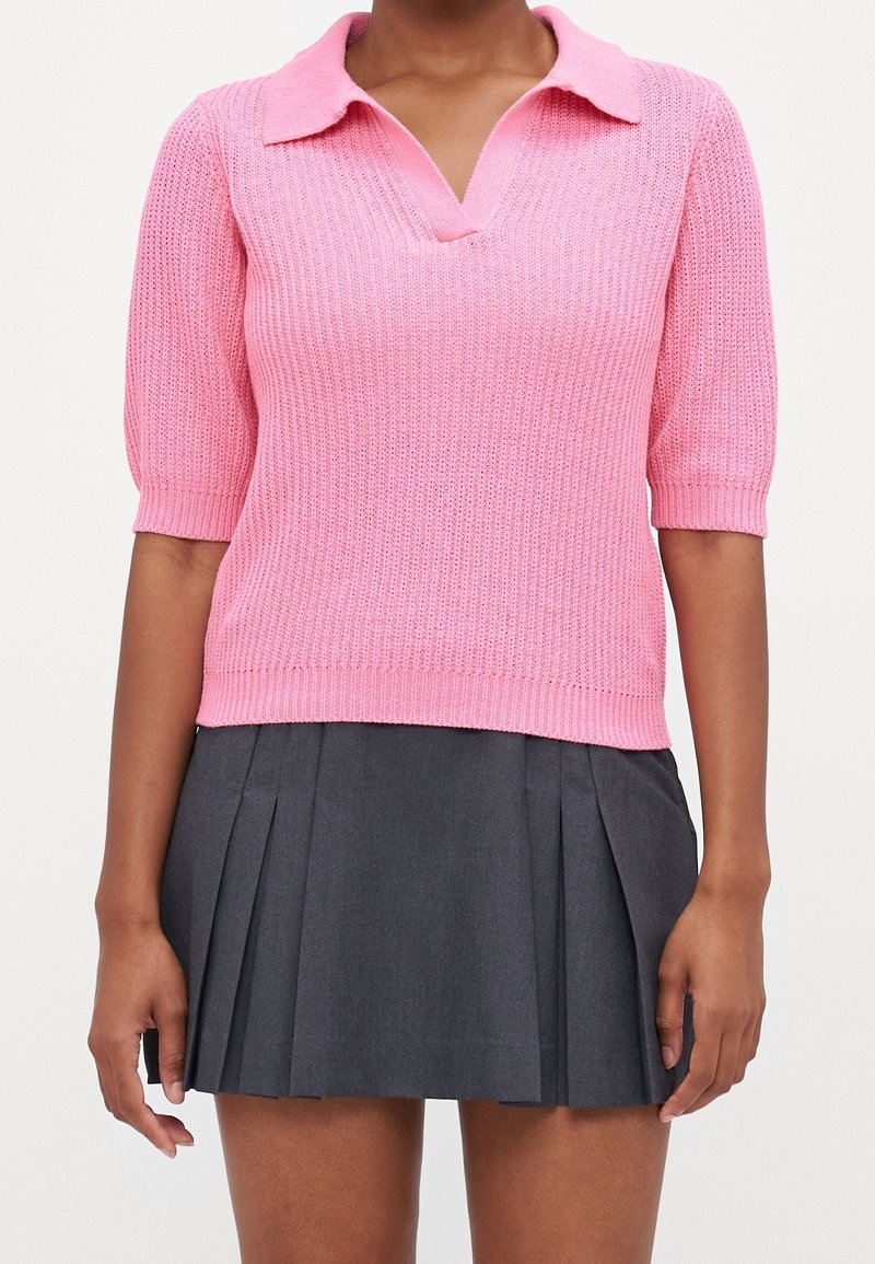 Personne portant un haut en tricot rose à manches courtes avec un col et une mini-jupe plissée gris foncé, debout devant un fond blanc.