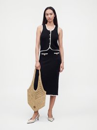 Femme portant un haut noir sans manches boutonné et une jupe avec un bord blanc, tenant un grand sac fourre-tout tressé beige et des chaussures blanches à talons.