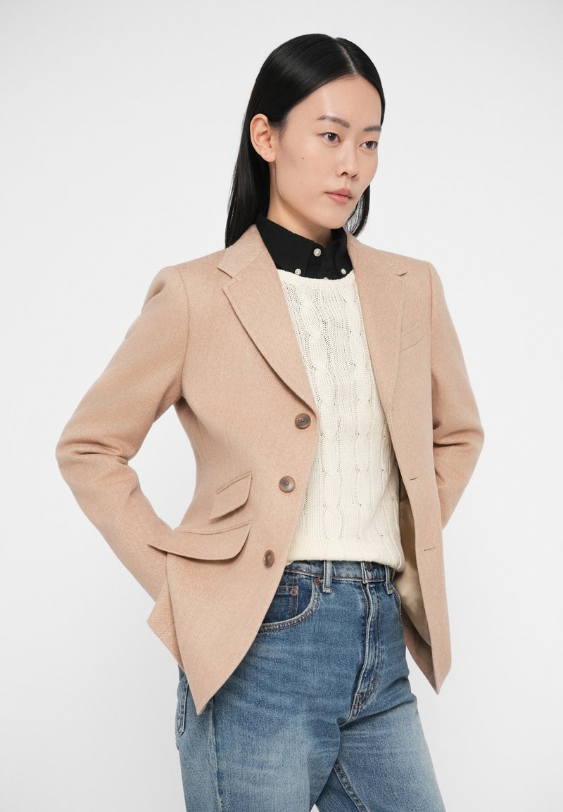 Blazer in lana rosa cipria con due bottoni, rever a lancia e tasche frontali, indossato sopra un maglione lavorato a trecce color crema e jeans blu.