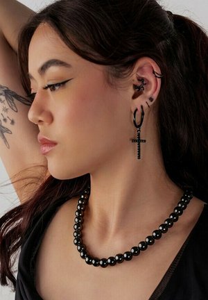 Junge Frau mit mehreren schwarzen Ohrpiercings, großem schwarzem Kreuzohrring, schwarzer Perlenkette und Wing- und Stern-Tattoo am Oberarm.